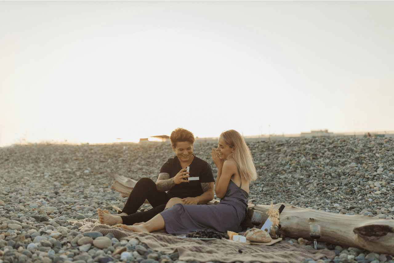A couple having a picnic on the beach, the man holding an open jewelry box and the woman smiling.