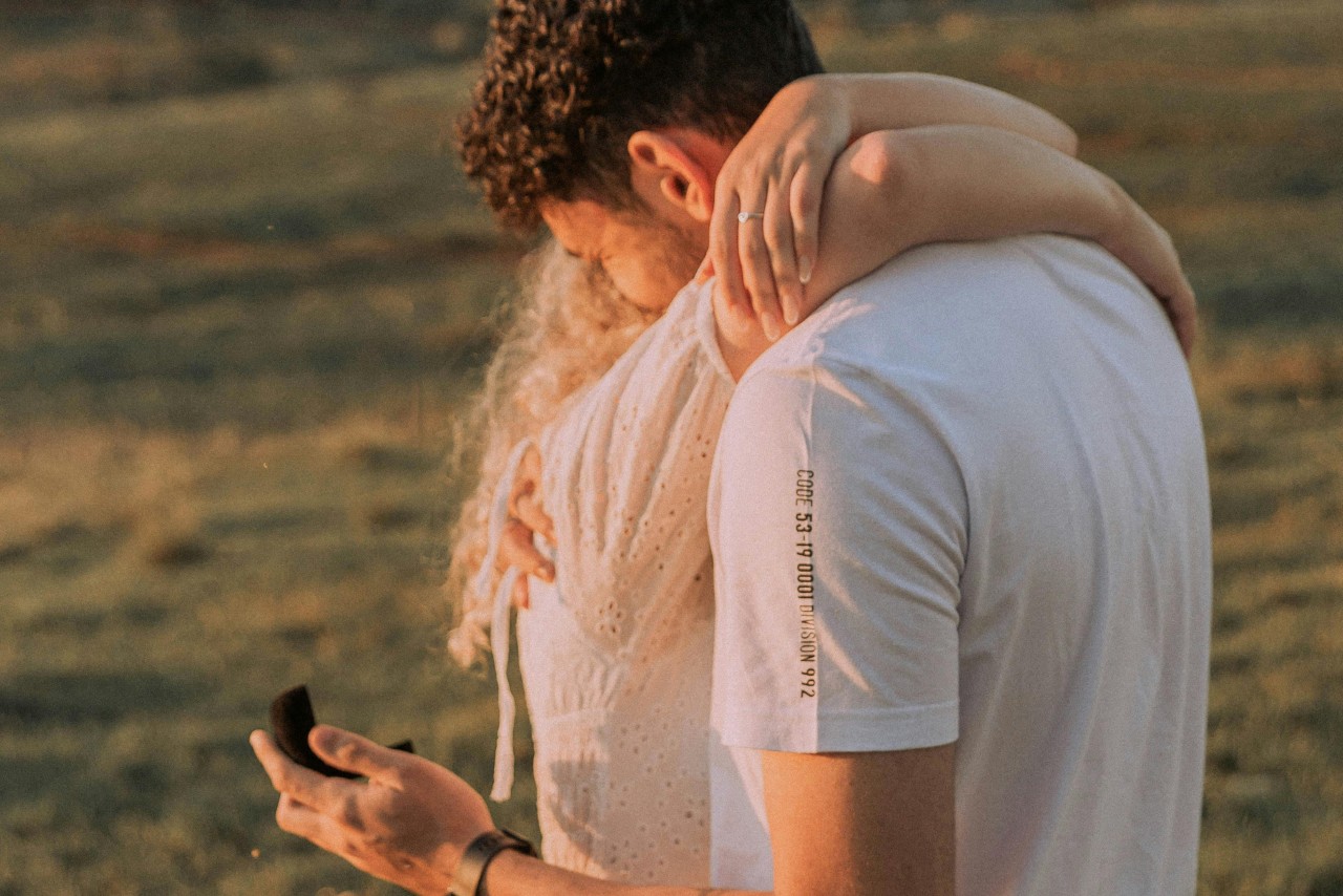 A woman embracing a man as he smiles and holds a ring box behind her back.