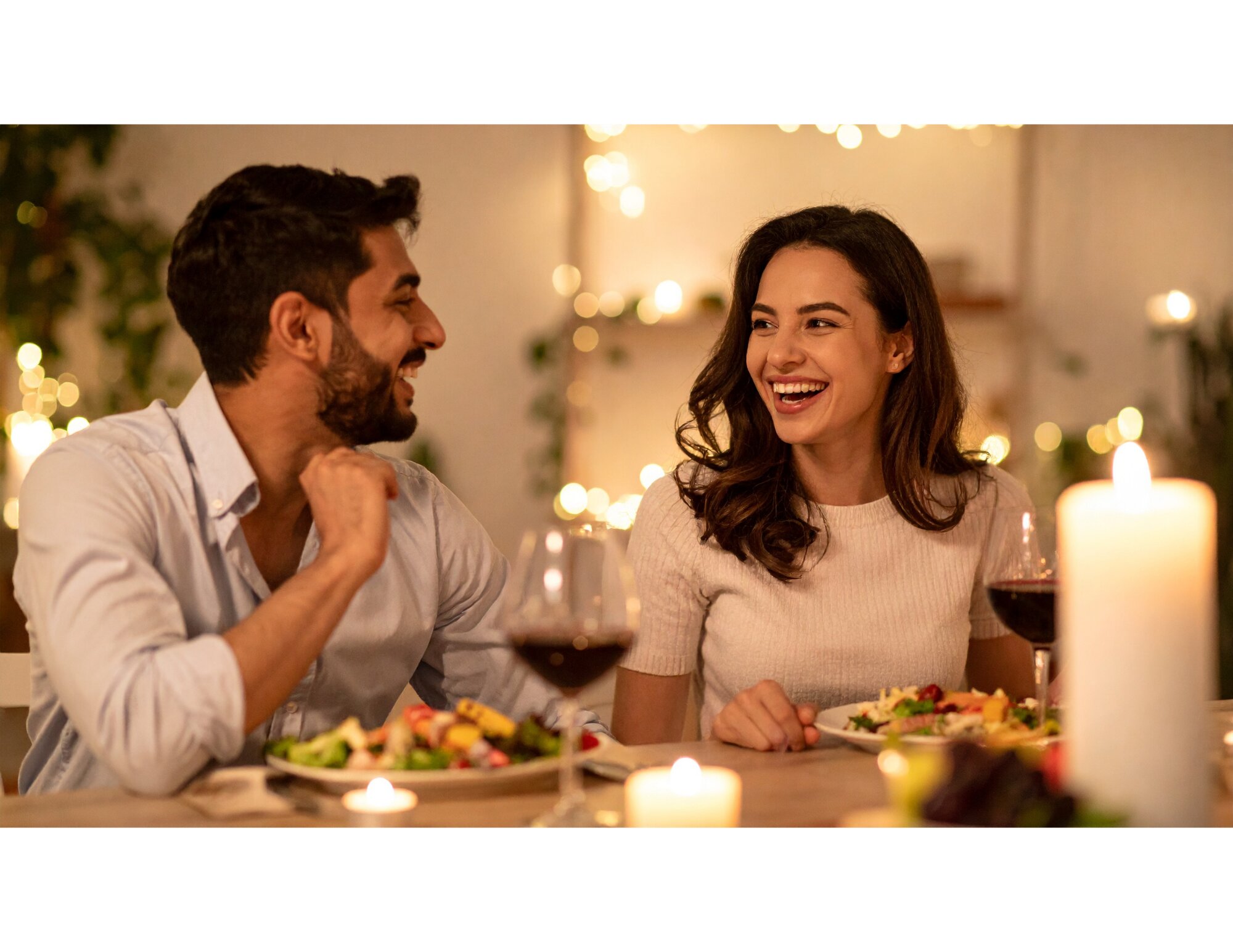 A couple is sitting at a table, the woman is smiling while enjoying a glass of wine and candlelit dinner, the man is wearing a nice dress shirt and smiling.