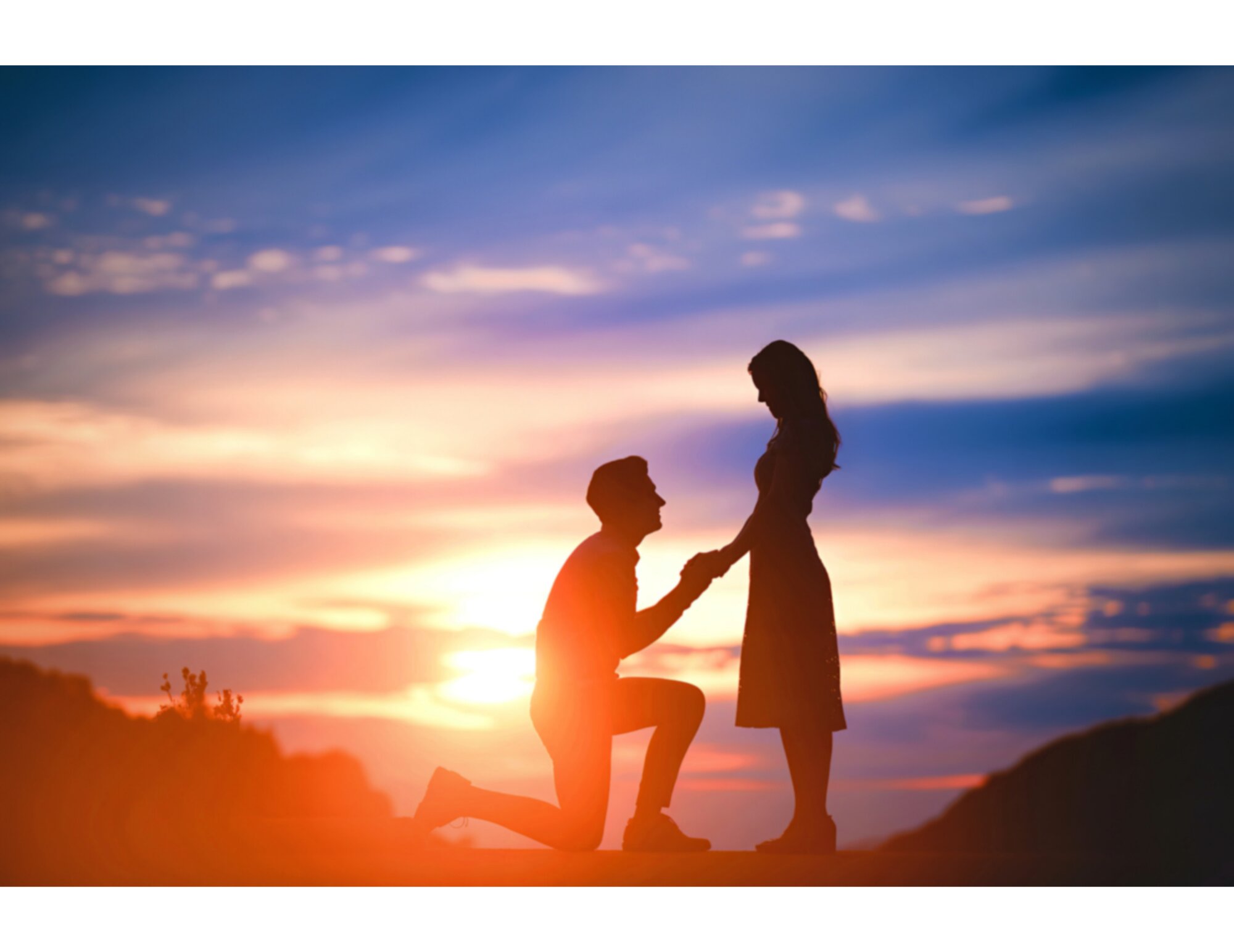Man kneeling on the sand holding a woman’s hand during a beach proposal with sunset in the background.