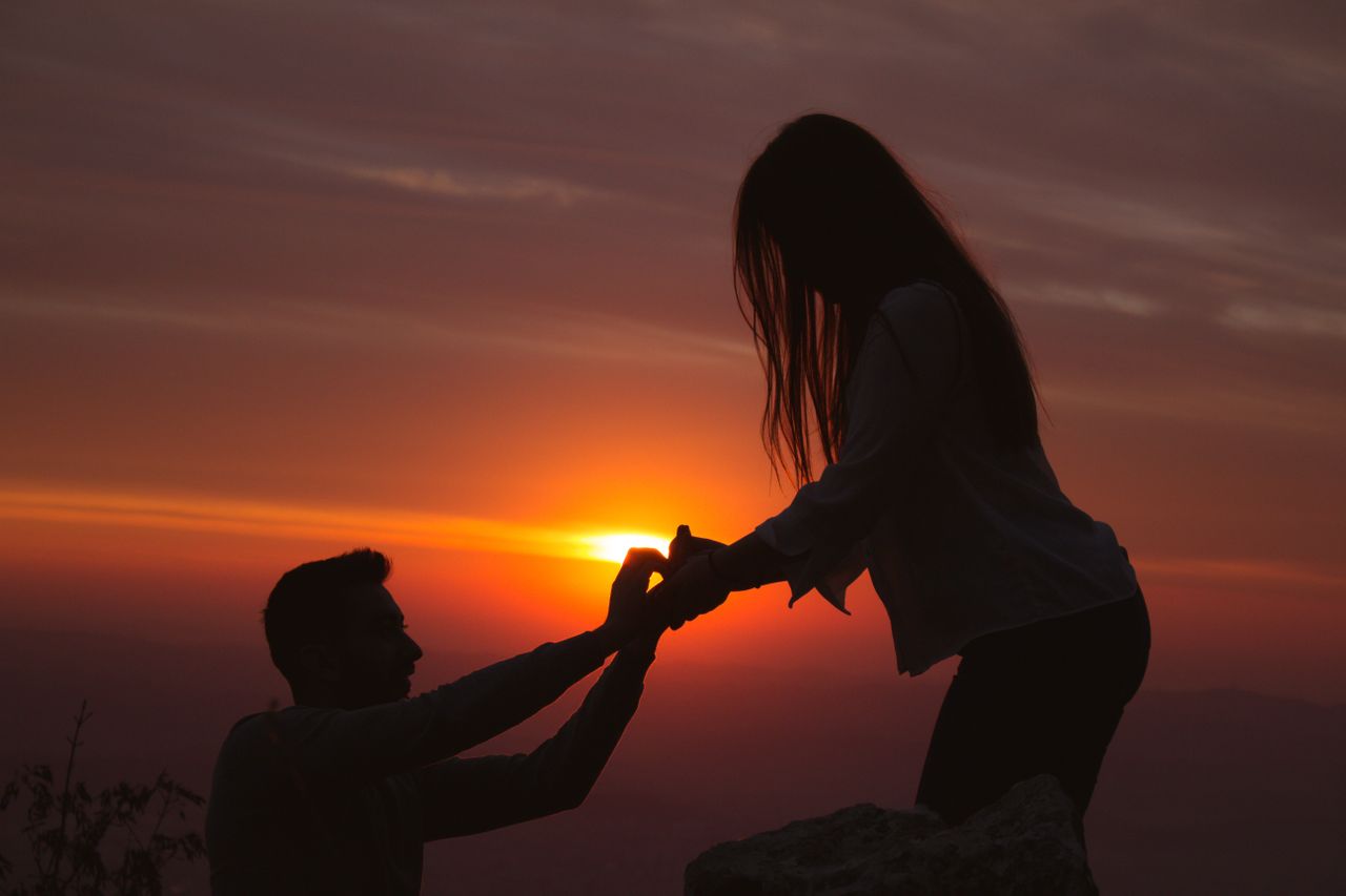 A man kneeling and proposing to his partner in front of the vibrant sunset.