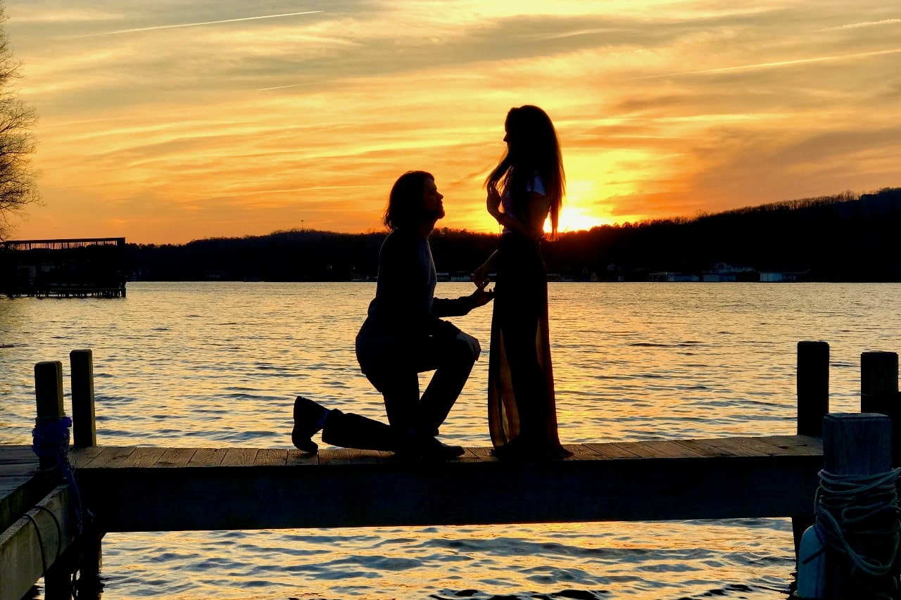 A man kneels to propose to a woman in front of a sunlight lake.