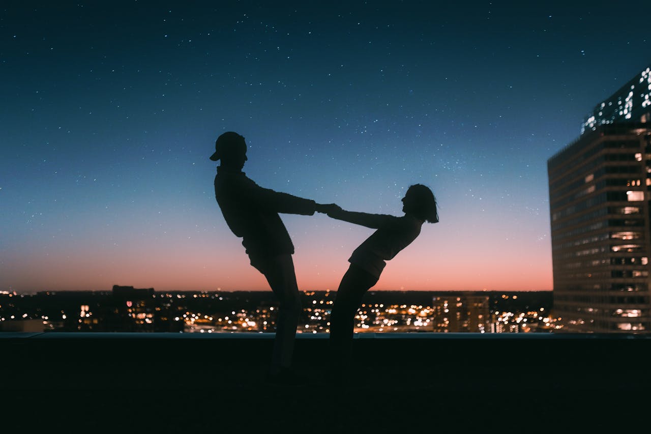 A couples holds each other hands and dances on a rooftop in front of a night sky full of stars.