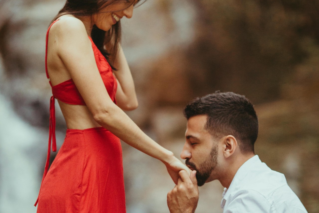A man in a white shirt is kneeled and kisses a woman’s hand in a red dress.