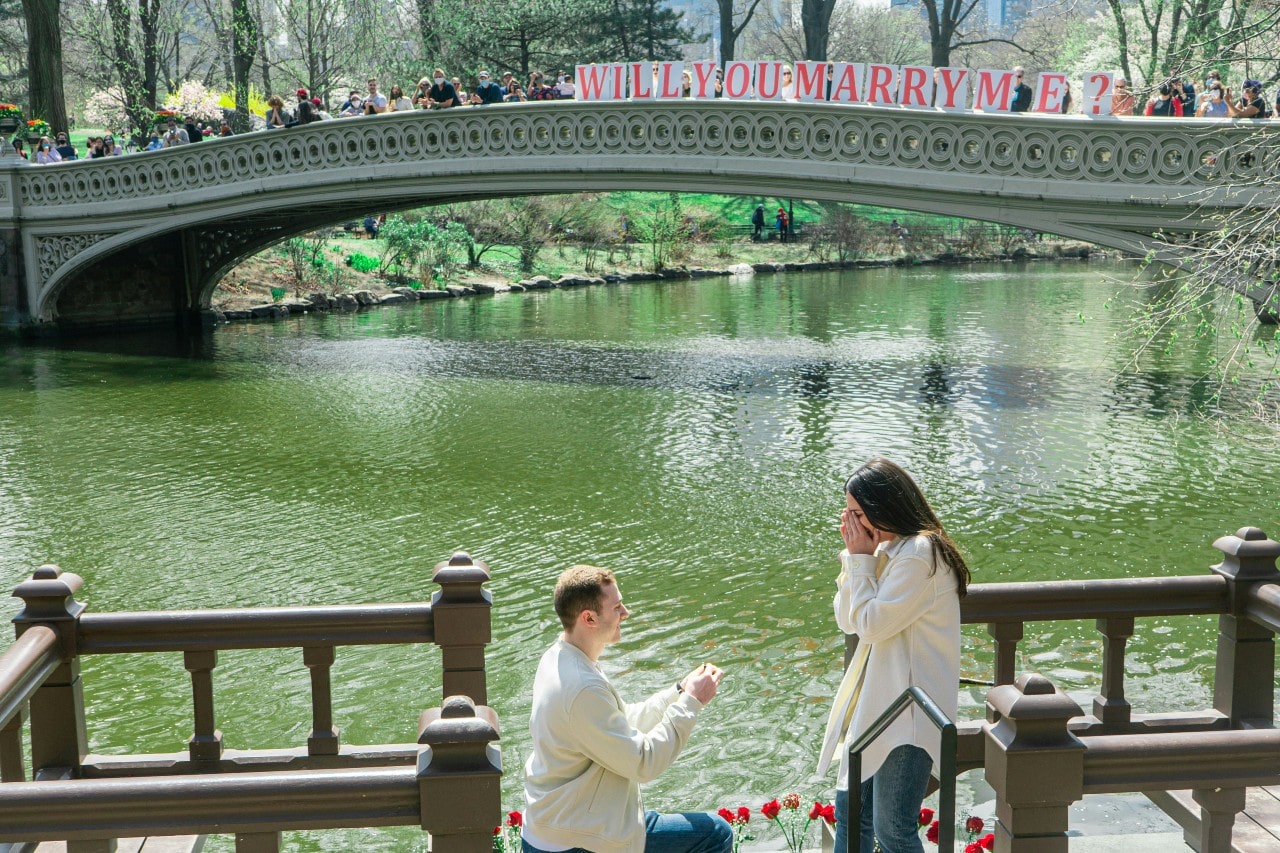 A man kneels to propose to a woman in front of a bridge, decorated with “WILL YOU MARRY ME” letters.o