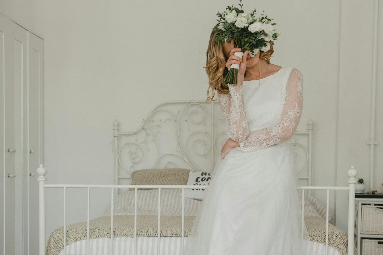 A bride in her gorgeous gown standing in front of a white vintage bed, a bouquet of flowers covering her face.