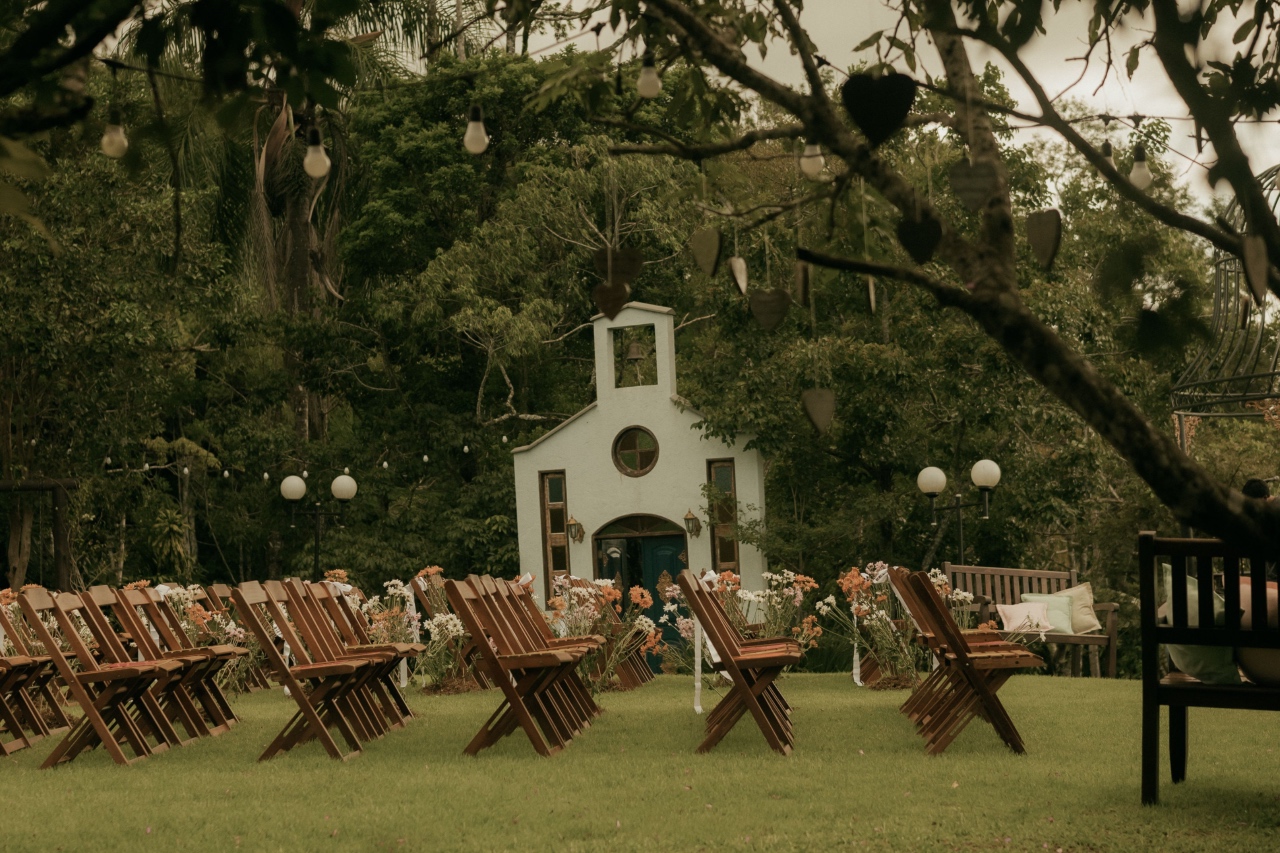 An outdoor wedding scene, filled with empty chairs and trees and a church in the background.