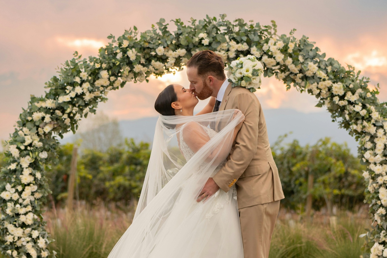 A newlywed couple kissing in front of a flower arc with a picturesque sunset in the background.