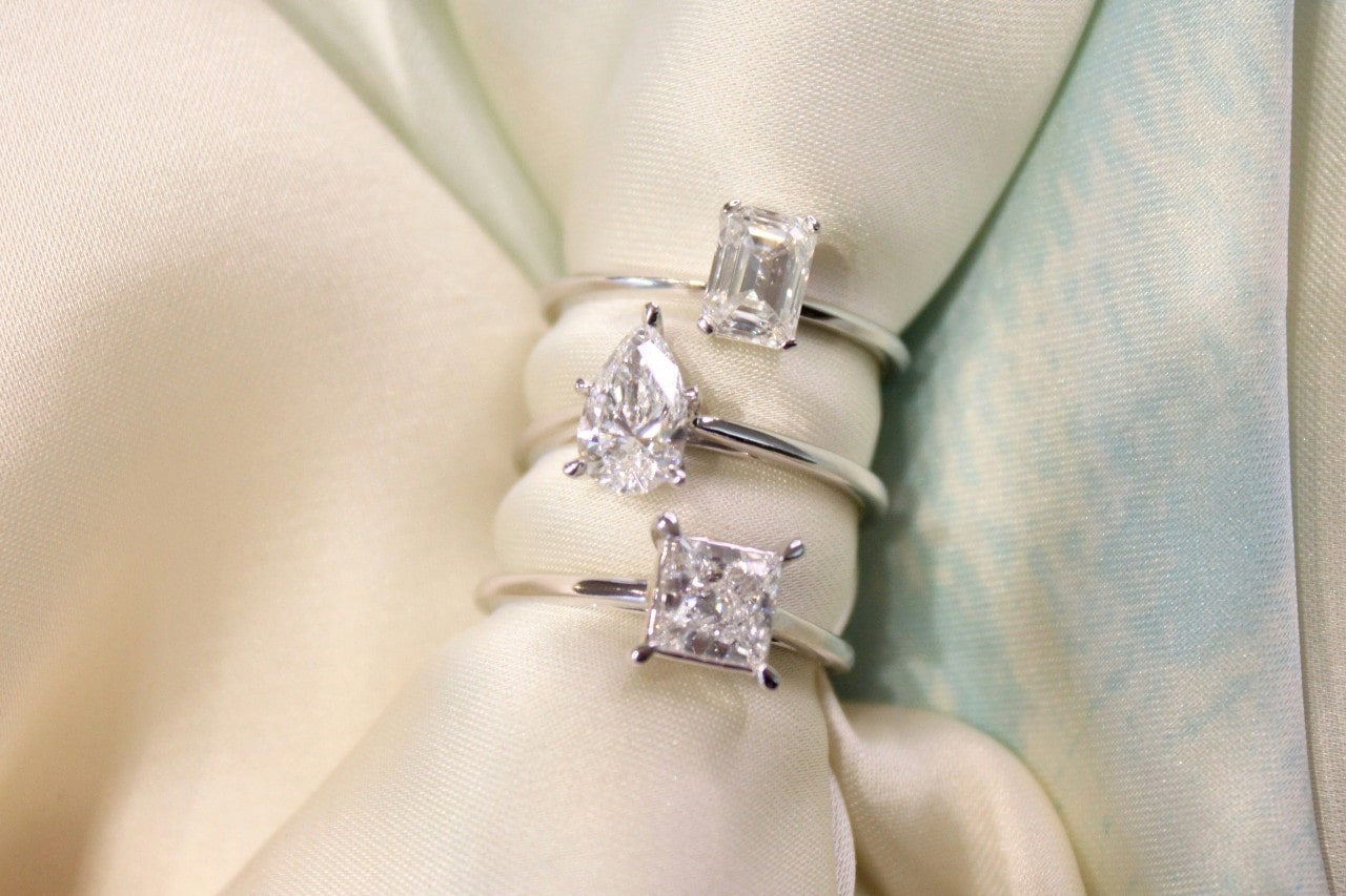 Three diamond engagement rings with different cuts (emerald, pear, and princess) displayed on satin fabric.