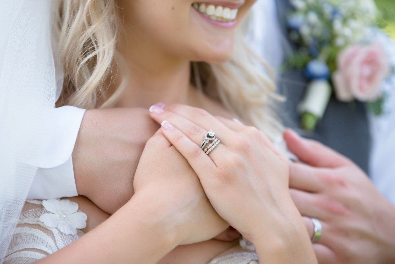A close-up of a smiling bride being embraced by a groom, showcasing her side stone diamond engagement ring and wedding bands.