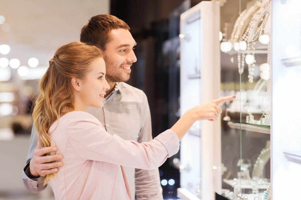 A happy couple window shopping at their local jewelry store, the woman pointing at a piece she loves.