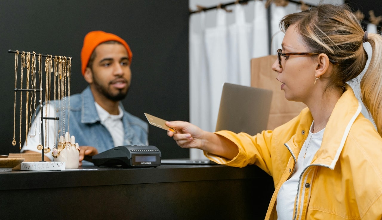 A woman in a yellow jacket taps a credit card on a terminal at a local jewelry store as she chats with the jeweler.