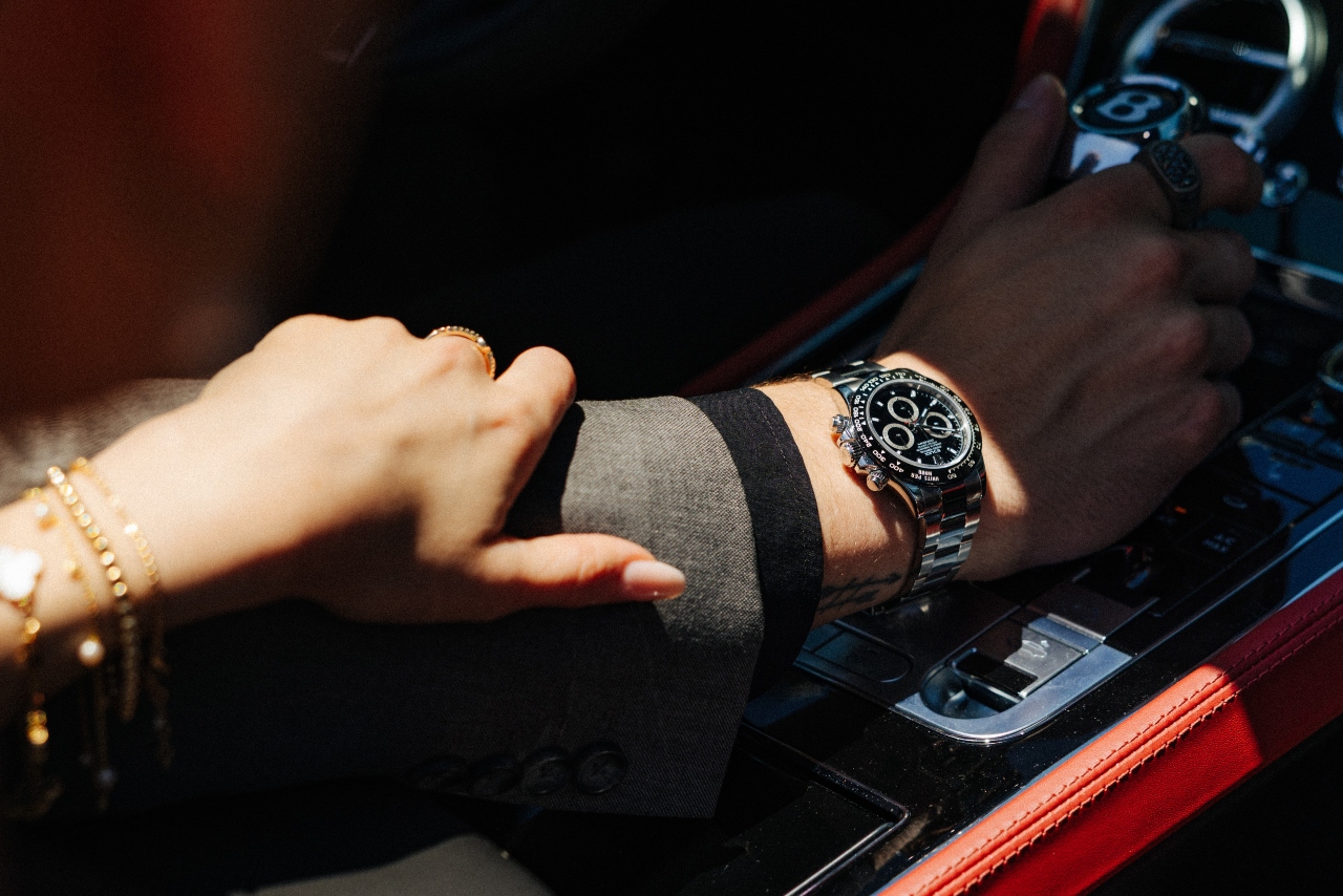 Close-up of a woman with gold bracelets holding onto a man wearing a luxury watch and driving a luxury sports car