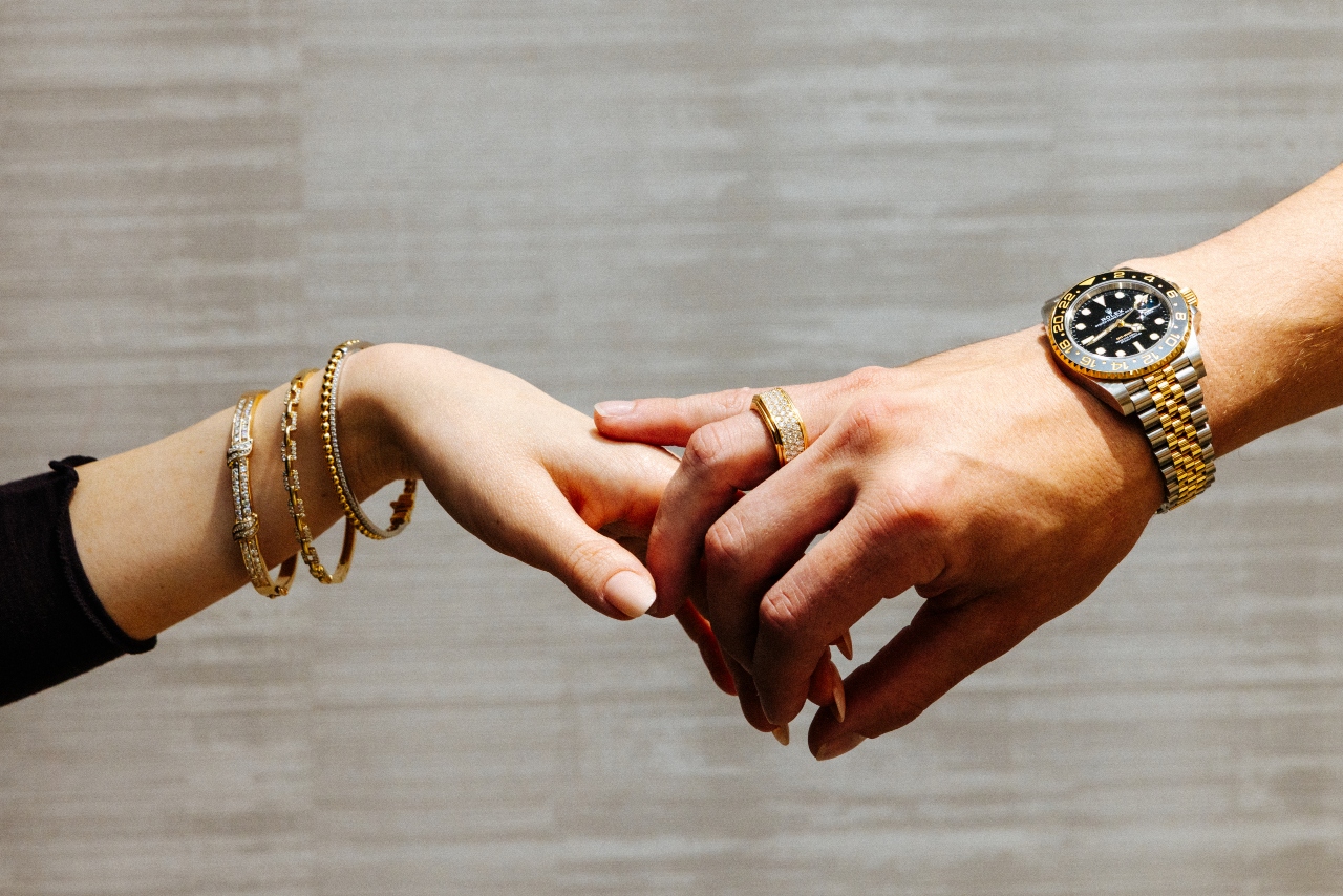 A man wearing a yellow gold multirow diamond ring and a stainless and yellow gold watch holding the hand of a woman with yellow gold and diamond bangles
