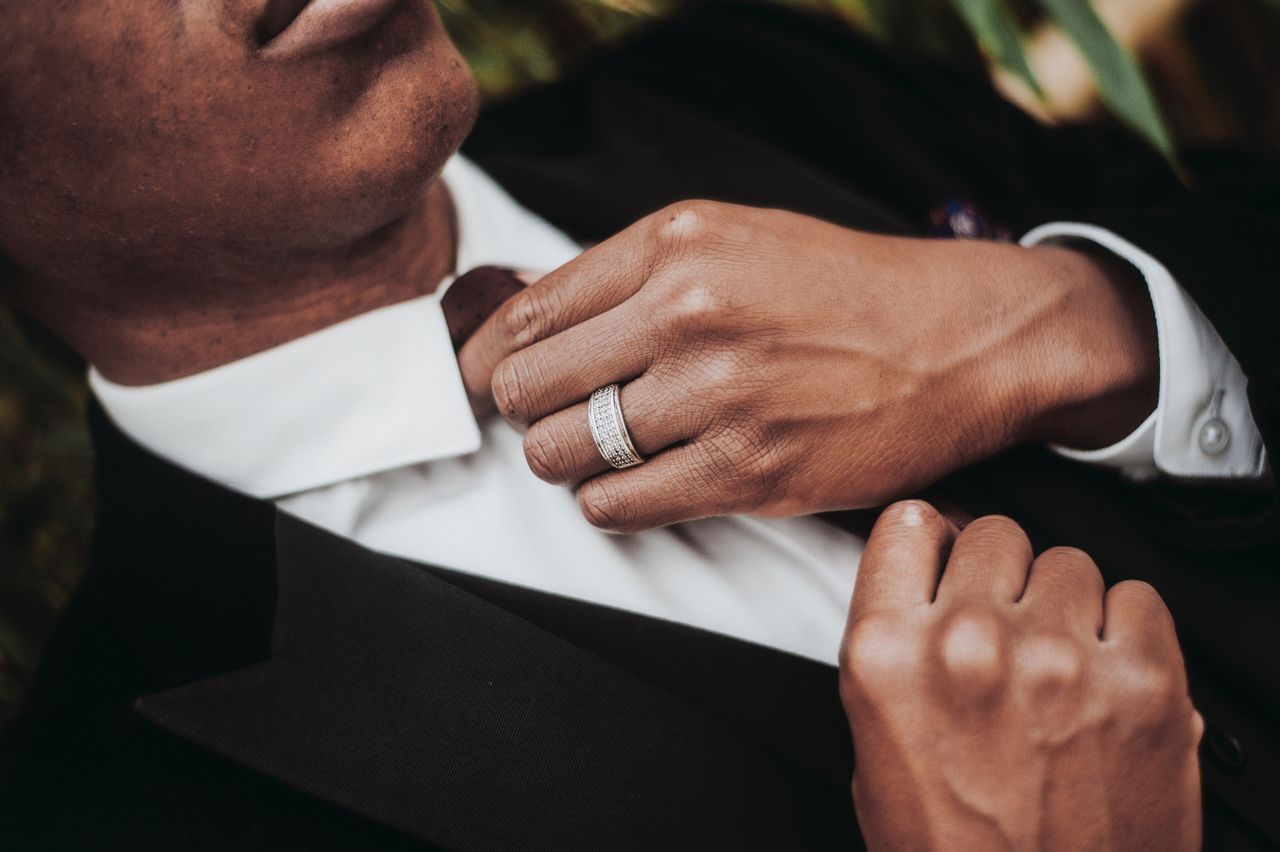 A close-up of a man wearing a textured silver-toned ring and a black suit adjusting his tie. A close-up of a man wearing a textured silver-toned ring and a black suit adjusting his tie.