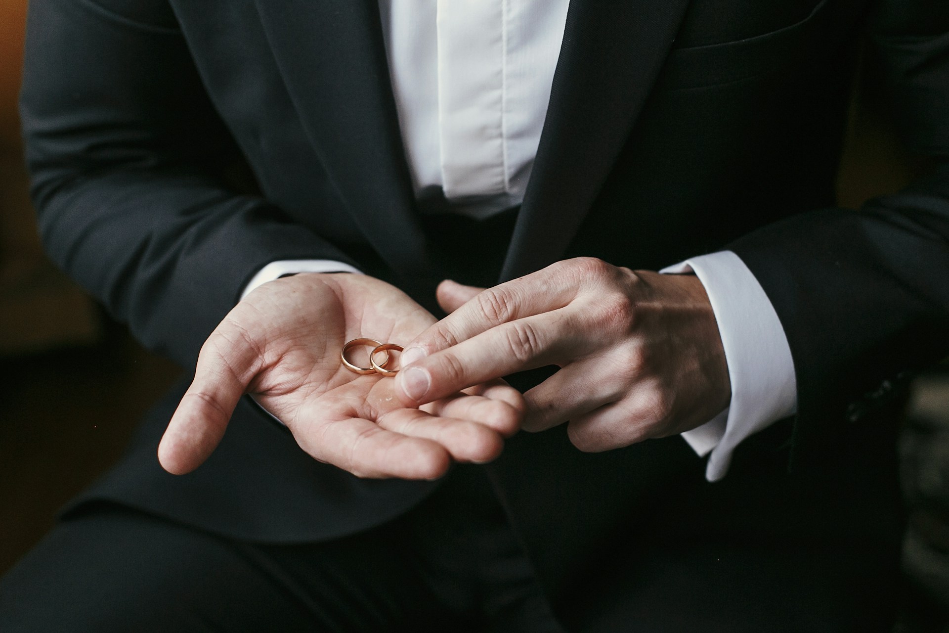 A close-up of a man in a dark suit holding two gold wedding rings in his palm. A close-up of a man in a dark suit holding two gold wedding rings in his palm.