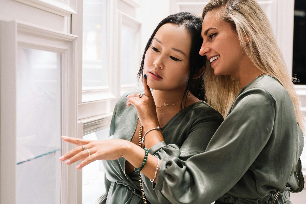 Two women in olive satin blouses look through a jewelry showcase.