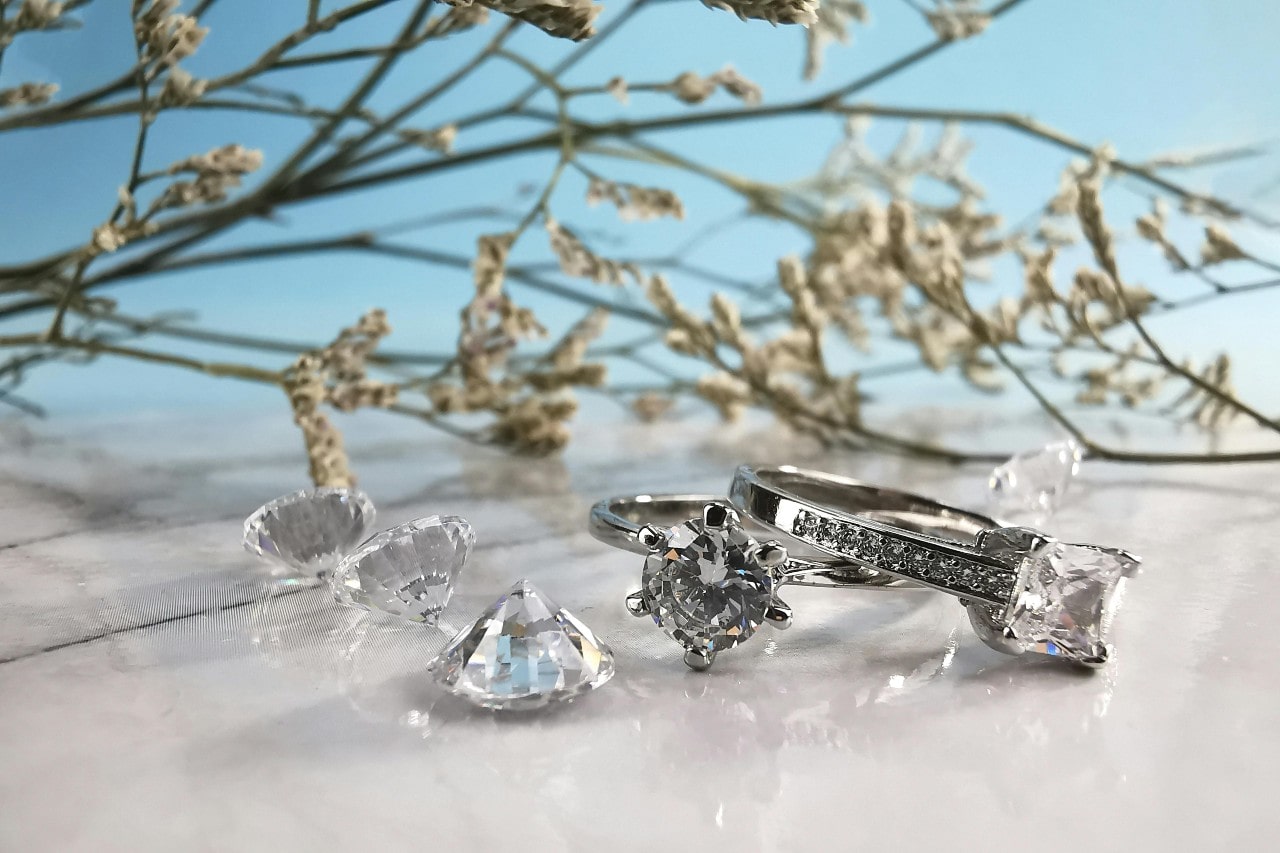 Two engagement rings and miscellaneous diamonds lying on a pearly white surface with a tree in the background. Two engagement rings and miscellaneous diamonds lying on a pearly white surface with a tree in the background.