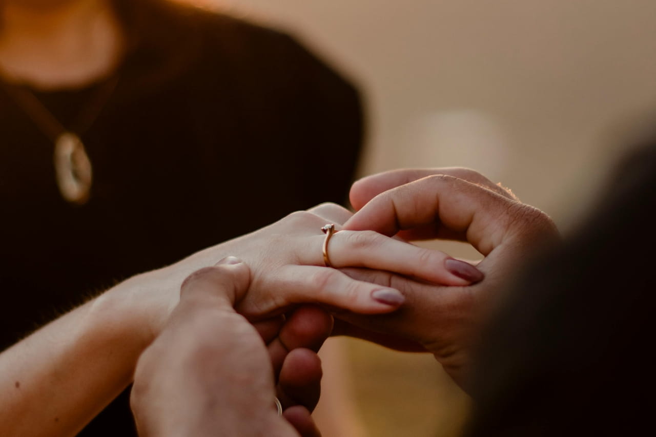 Close-up of the couple's hands as a man places a solitaire ring on the woman's finger, against a warm, blurred background. Close-up of the couple's hands as a man places a solitaire ring on the woman's finger, against a warm, blurred background.