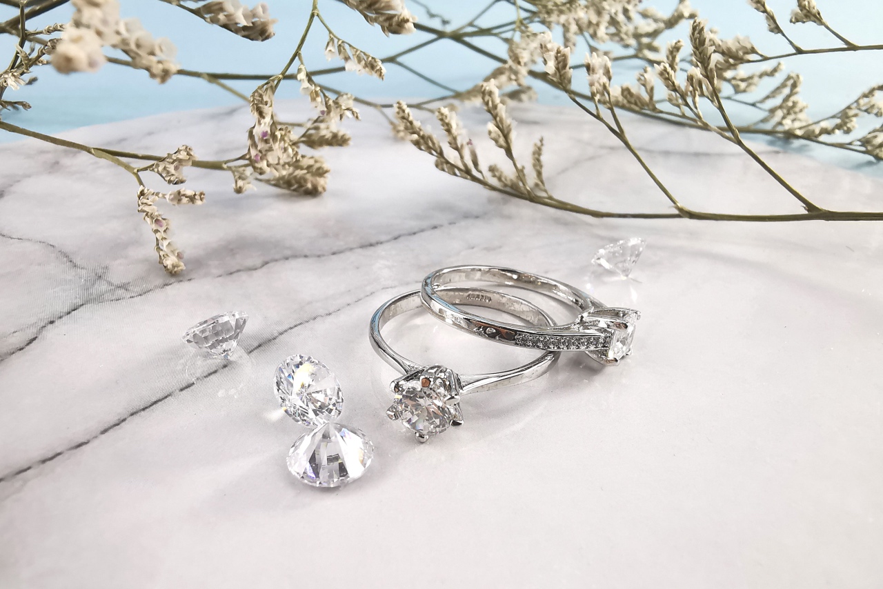Two white gold diamond engagement rings displayed on marble next to loose diamonds and dried flowers.