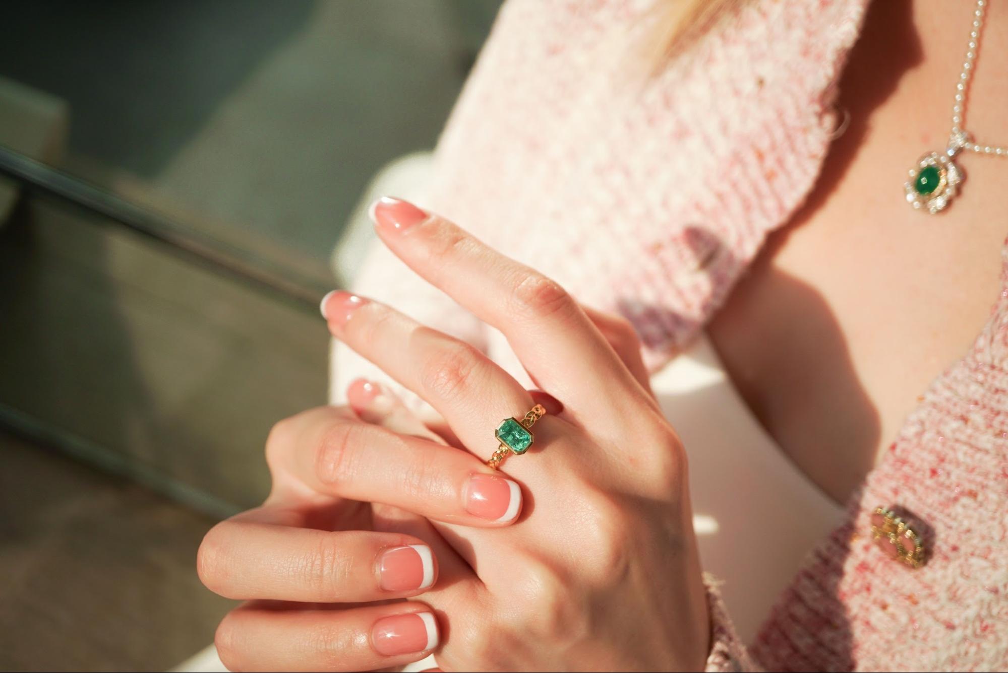 A close-up of a woman's hand with a French manicure wearing a gold ring and a necklace with an emerald stone.