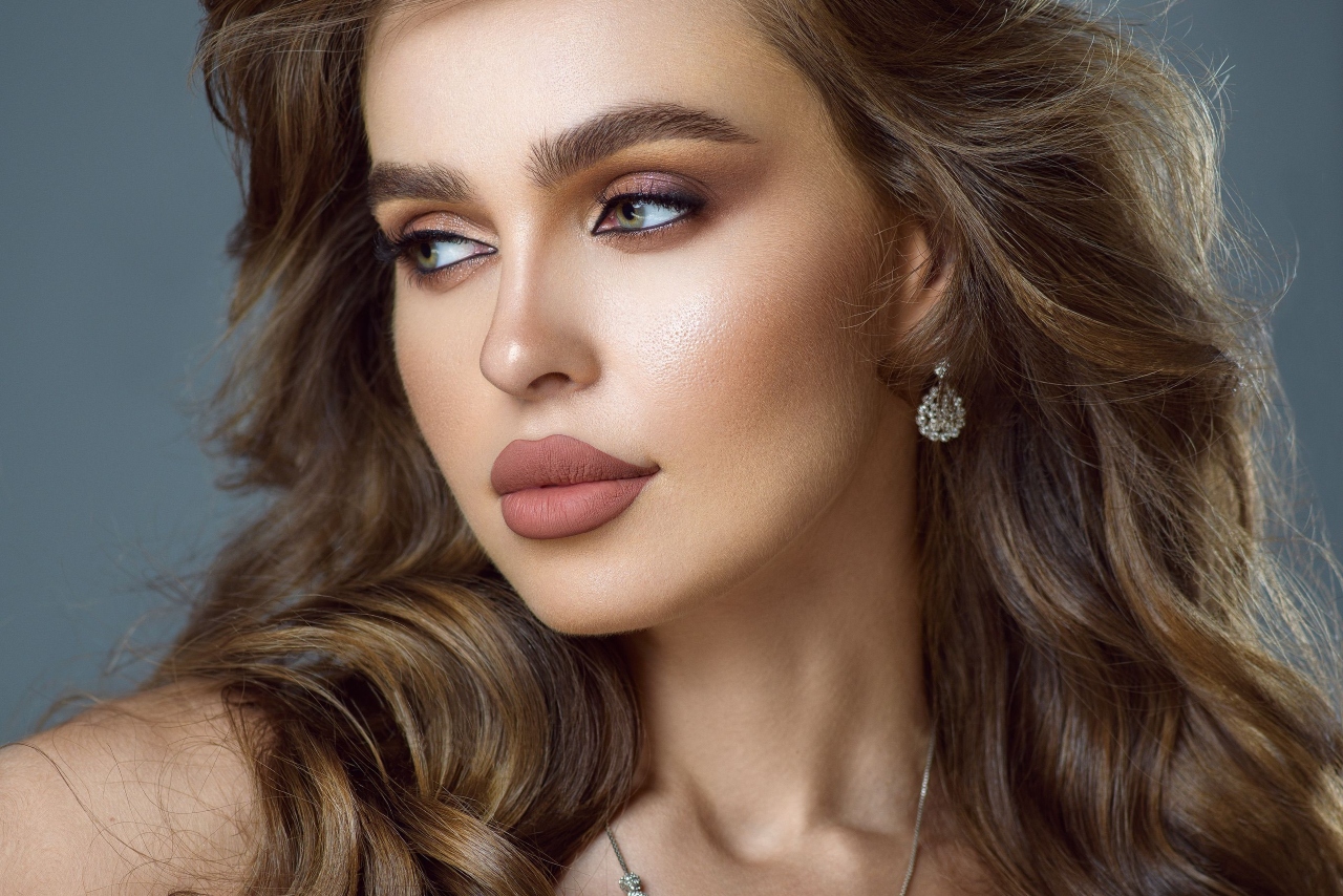 A close-up of a woman with glamorous makeup, wavy brown hair, and diamond drop earrings.