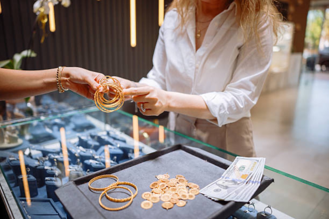A customer hands gold bangles to a jeweler over a glass display case holding gold coins and cash.