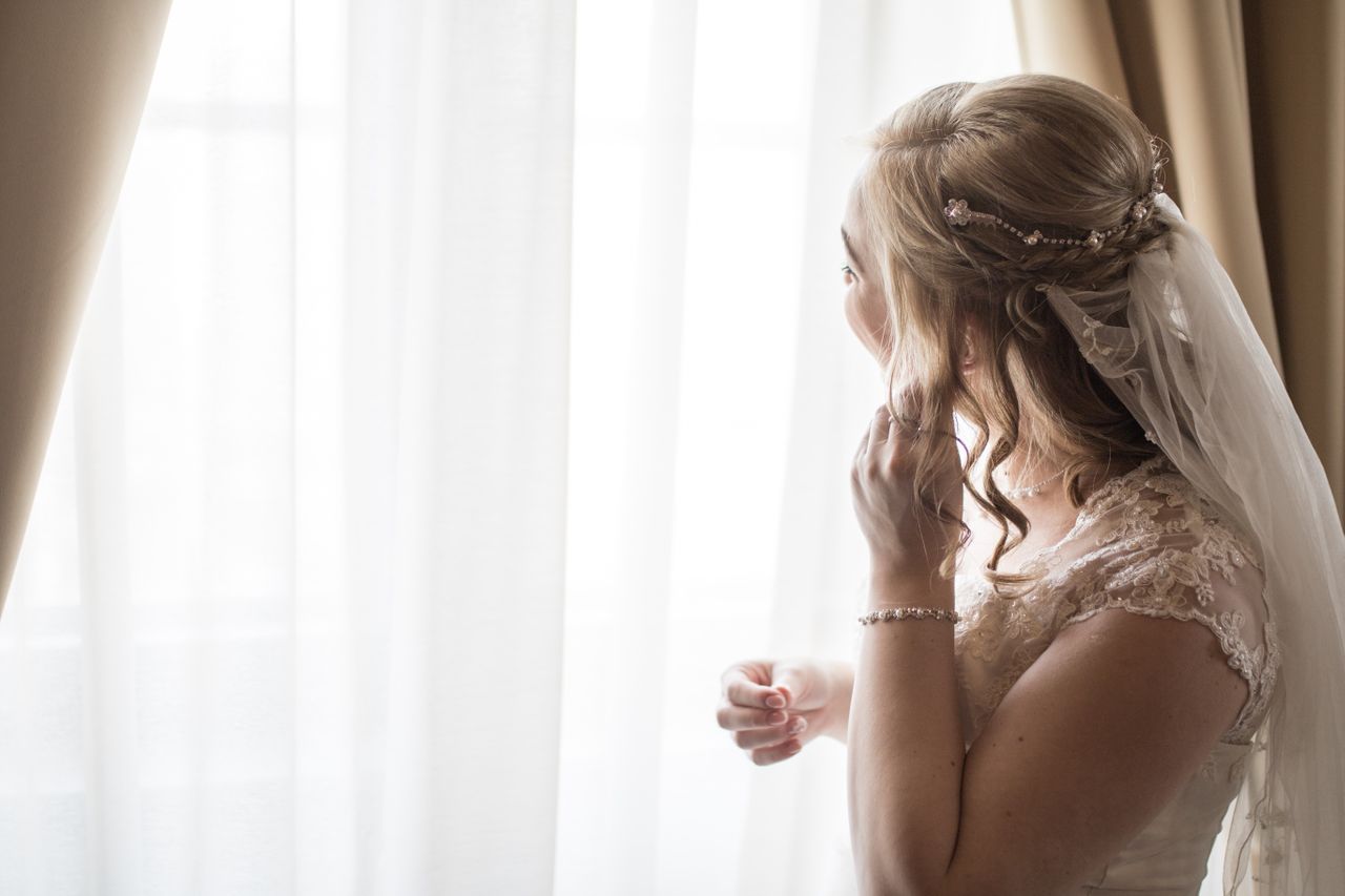 A bride on her wedding day touching her earring while staring out the window.