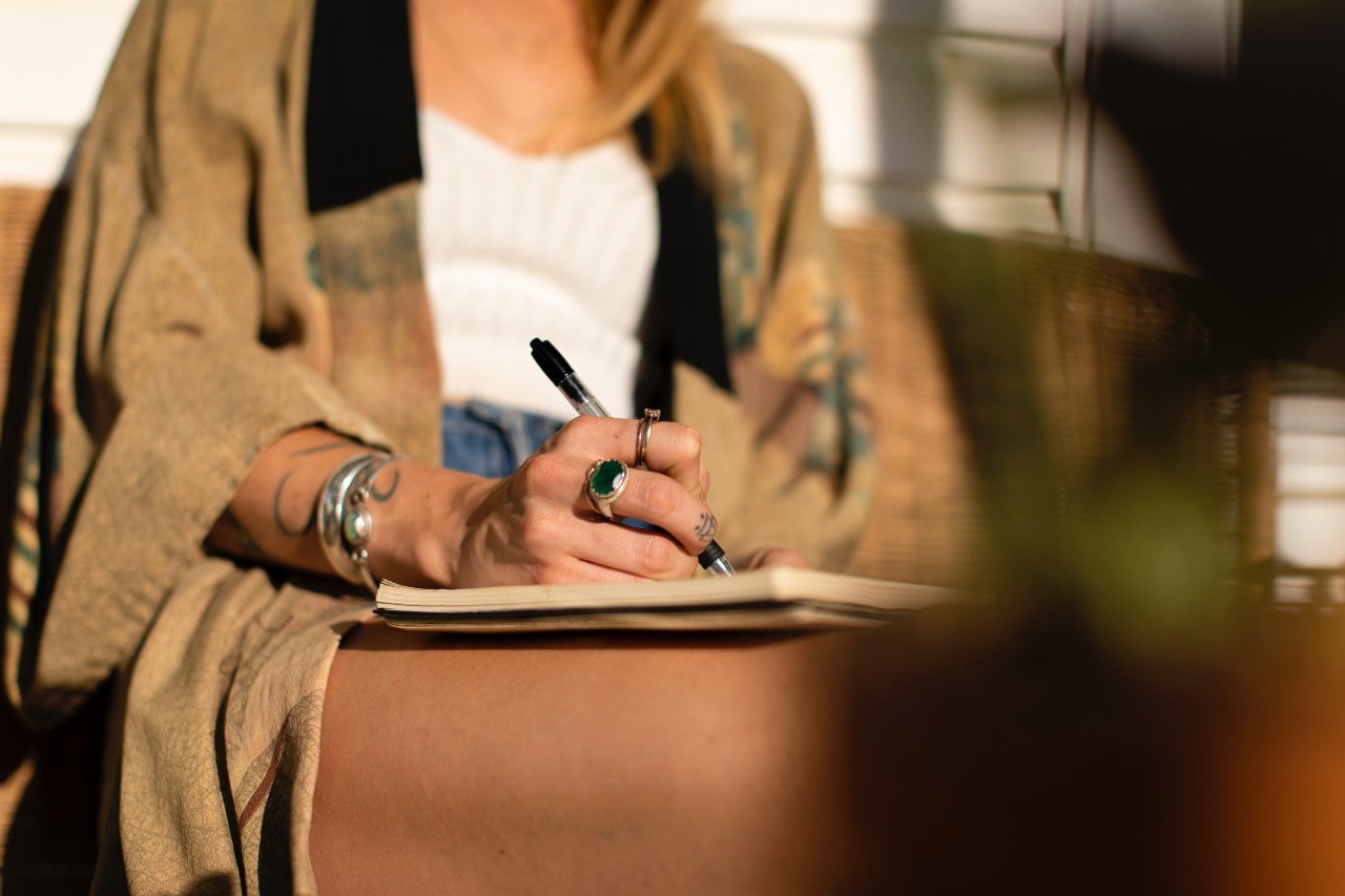 A woman writing in a notebook while sitting outside, her hand adorned with a large emerald ring and her wrist adorned with a silver bangle.